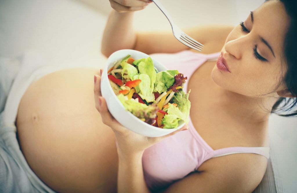 Closeup topview of late 20's pregnant woman eating a salad. She's sitting on sofa in her living room. The salad is in a white round bowl. Her 8 months round belly is visible. She's wearing pink sleeveless tanktop and beige pants. Toned image.